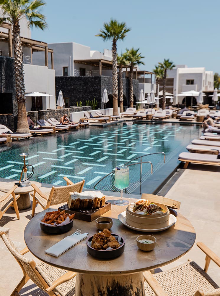 A poolside table at a luxurious resort with a variety of dishes, including bao buns, sweet potato fries, and chicken bites, accompanied by a blue cocktail, set against the backdrop of a stylish pool, lounge chairs, and palm trees.