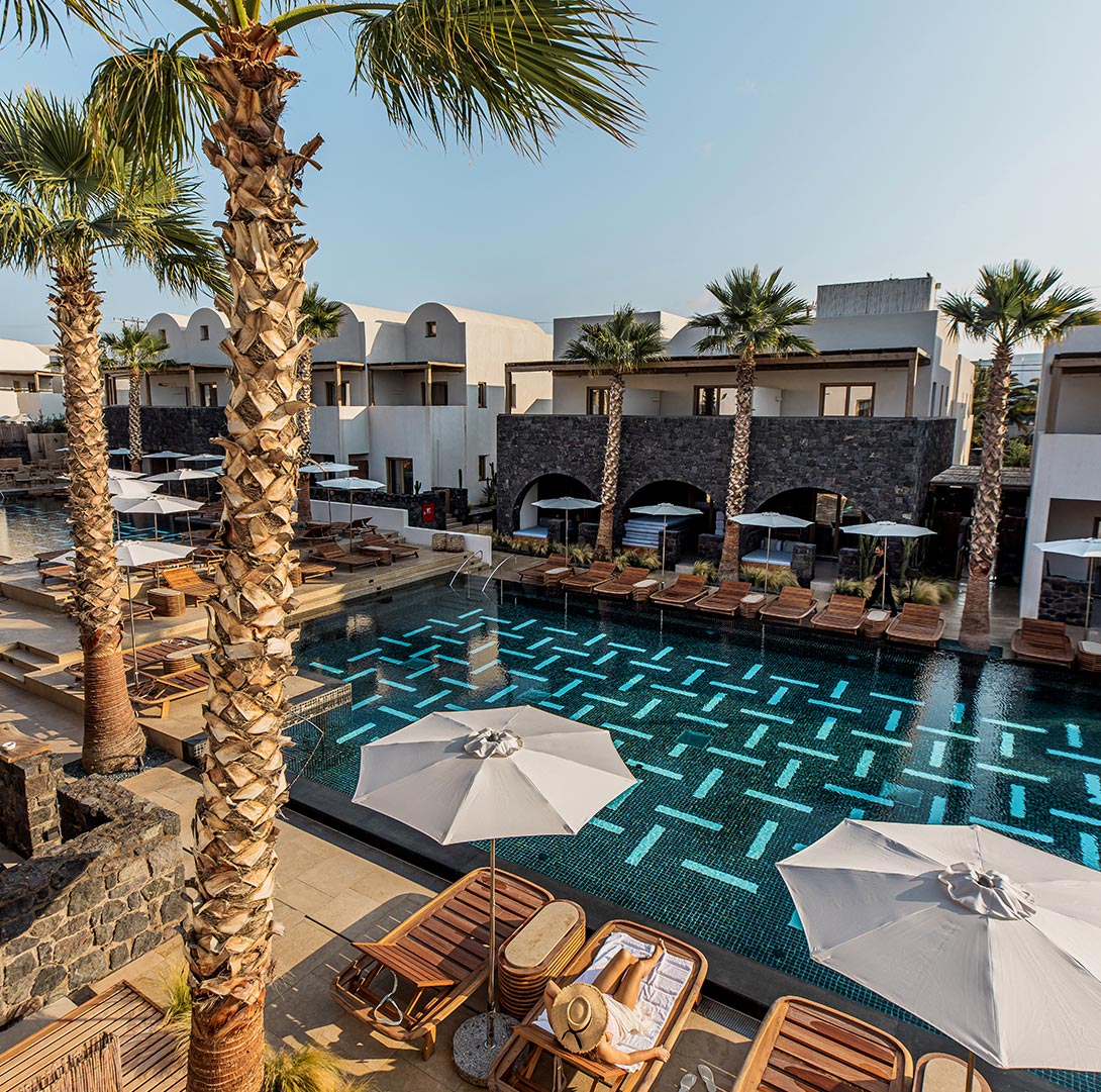 A luxurious resort featuring a modern pool with geometric tile patterns, surrounded by wooden lounge chairs, umbrellas, and palm trees, with whitewashed and stone-accented buildings in the background.