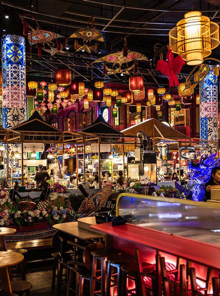 This image shows a vibrant indoor market scene with colorful hanging lanterns, ornate umbrellas, and traditional Thai decor. The foreground features a seating area with wooden tables and chairs, while the background is bustling with food stalls and floral arrangements, creating an atmosphere reminiscent of a Thai street market.