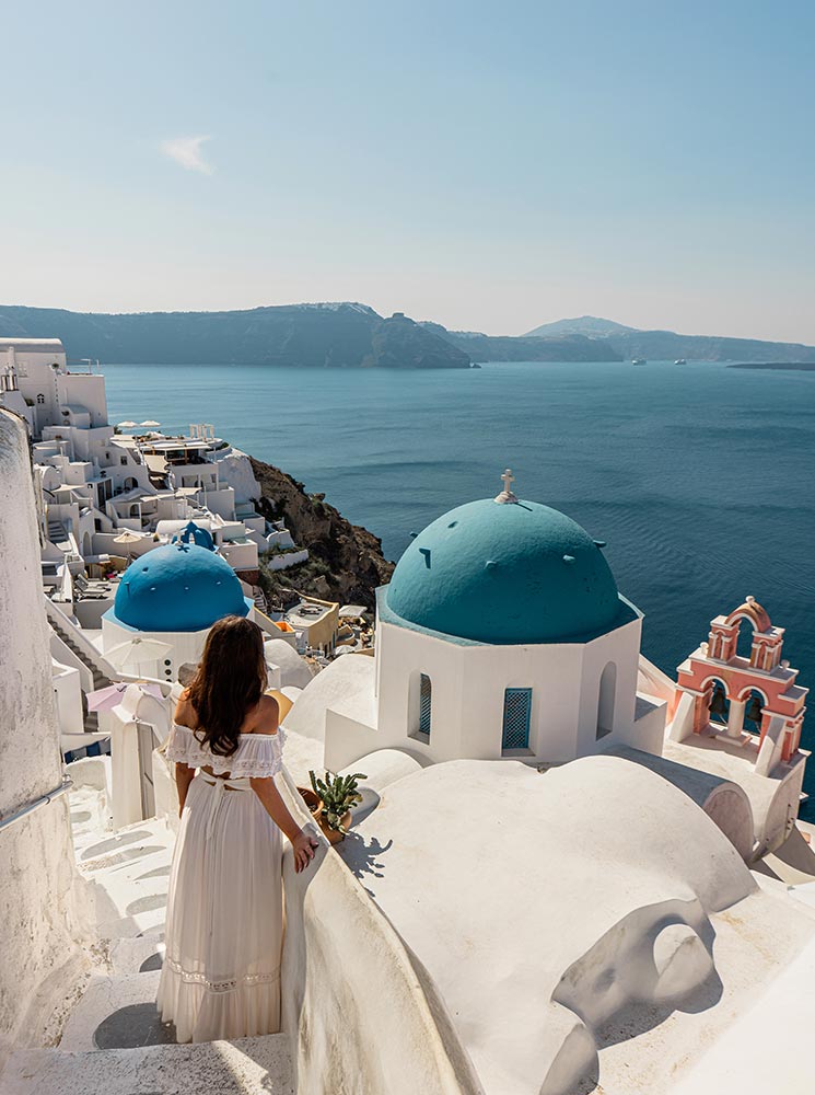 A woman in a white dress standing on steps overlooking the iconic blue-domed churches of Oia, Santorini, with whitewashed buildings and the deep blue Aegean Sea stretching into the horizon.