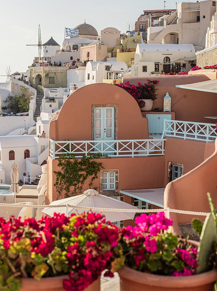 A picturesque view of Oia, Santorini, featuring colorful houses with blue balconies, blooming bougainvillea flowers, and whitewashed buildings under the warm Mediterranean sunlight, with a Greek flag waving in the background.