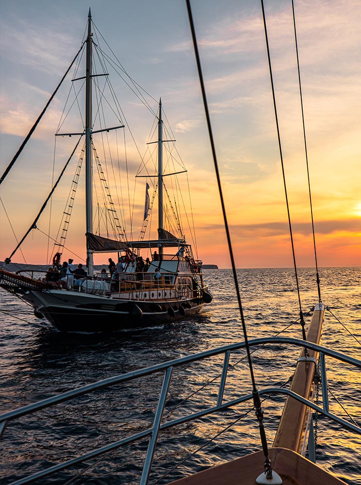 A picturesque scene of a traditional sailboat on the water during a Santorini sunset cruise, with vibrant orange and pink hues in the sky reflecting on the calm sea.