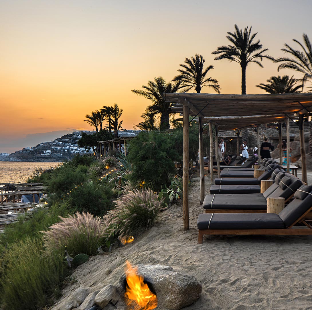 A serene view of Scorpio’s Beach Club in Mykonos at sunset, featuring shaded wooden lounge chairs, palm trees, and a sandy path adorned with lush greenery and a small fire pit, overlooking the calm sea and a softly lit horizon.