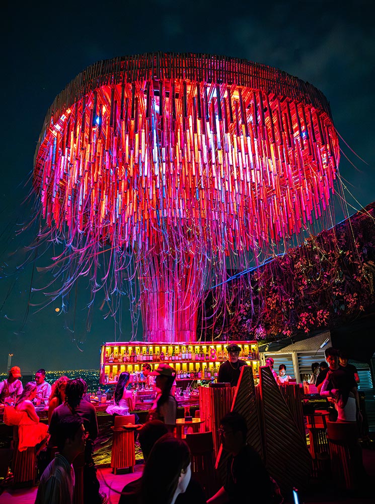 This image features a stunning rooftop bar illuminated by a vibrant, tree-shaped light installation glowing in shades of pink, red, and purple. Patrons gather around the bar, which is lined with shelves of colorful bottles, creating a lively and modern atmosphere. The dark night sky and the city lights in the background further enhance the dramatic and upscale ambiance of the setting.