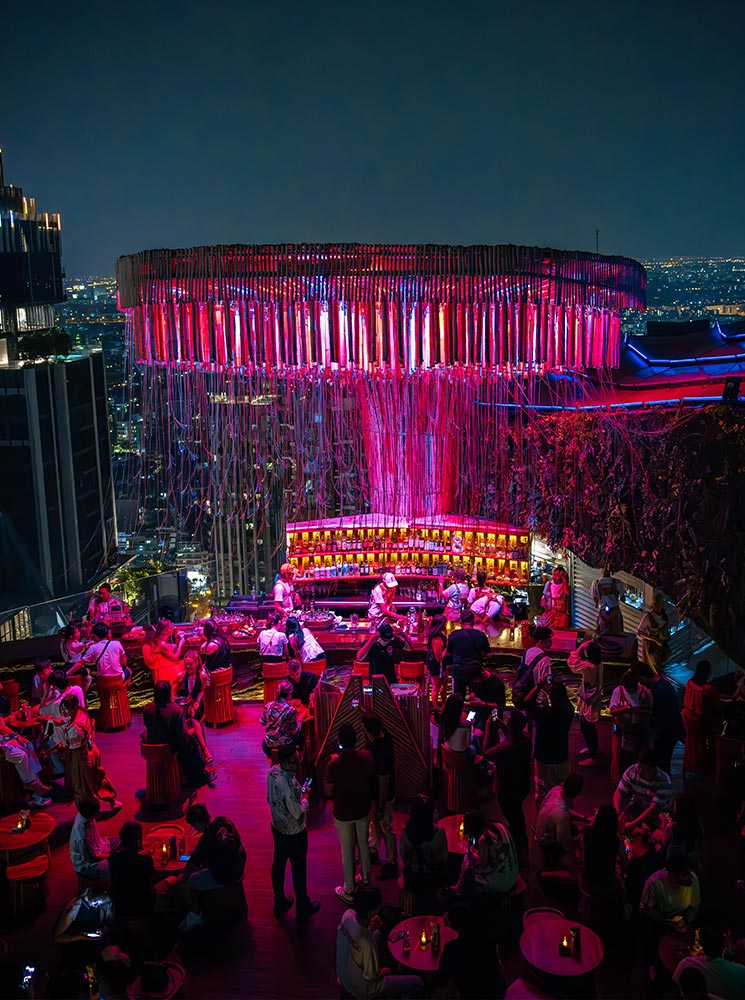 This image showcases a vibrant rooftop bar in Bangkok at night, featuring a colorful circular centerpiece illuminated with pink and purple lights. The lively atmosphere is enhanced by patrons seated around wooden tables and stools, enjoying drinks with a stunning cityscape in the background. The modern, neon-lit decor creates an energetic and stylish ambiance.