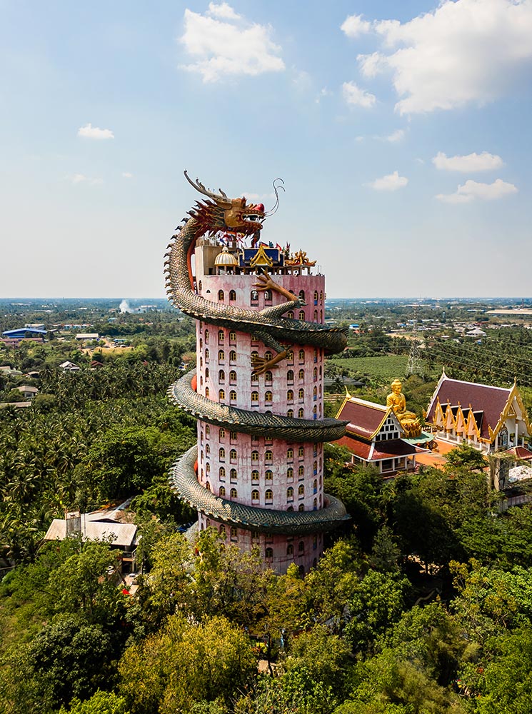 This image features Wat Sam Phran, a towering pink temple in Bangkok encircled by a massive green dragon sculpture that coils up the structure. Surrounded by lush greenery and a sprawling landscape, the temple stands out as a unique architectural marvel, with additional traditional temple buildings and a golden Buddha statue visible in the background.