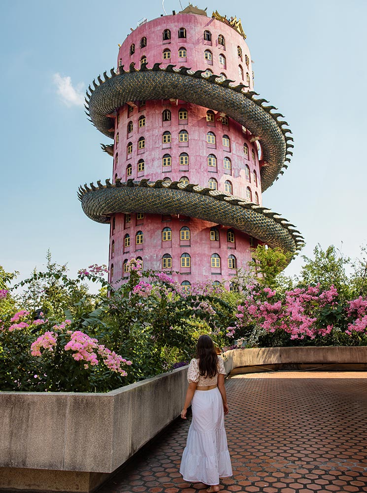 This image showcases the unique Wat Sam Phran temple in Bangkok, a striking pink cylindrical building wrapped by a massive green dragon sculpture. The foreground features a woman in a white dress walking toward the temple, surrounded by lush greenery and blooming pink flowers, creating a serene and awe-inspiring scene.