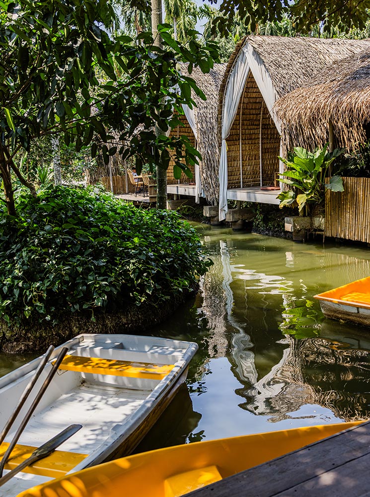 This image features a serene canal lined with tall palm trees and lush greenery, creating a tropical oasis. A woman in a white dress is paddling a yellow boat, adding a peaceful and picturesque element to the tranquil scene. The vibrant blue sky above enhances the natural beauty of this idyllic setting.