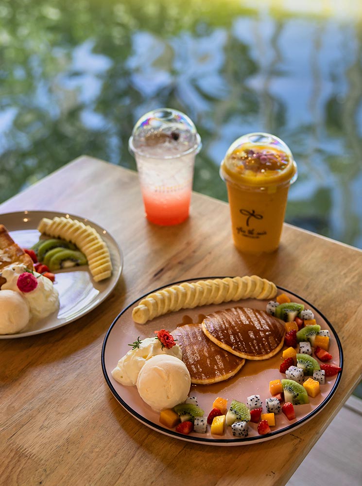 This image features a beautifully arranged plate of pancakes topped with syrup, served alongside sliced banana, colorful fresh fruit like dragon fruit, kiwi, mango, and strawberries, and scoops of vanilla ice cream. In the background are two refreshing drinks—one pink and one orange—with a blurred view of water reflecting sunlight, adding a serene ambiance to the cozy dining scene.