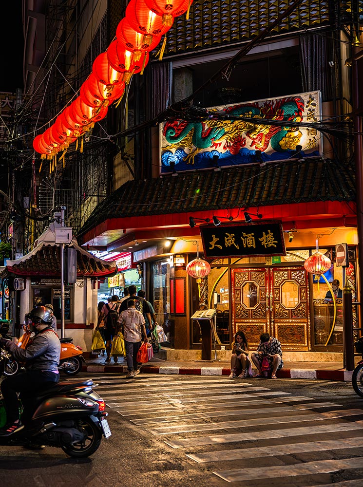 This image features a lively street scene in Bangkok’s Chinatown at night, with vibrant red lanterns hanging across the street and a richly decorated building with a dragon mural above its entrance. The warmly lit restaurant entrance, flanked by intricate Chinese-style doors and lanterns, adds to the inviting atmosphere. Scooters, pedestrians, and street activity complete the dynamic urban setting.