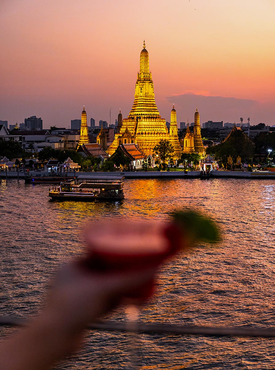 This image features the iconic Wat Arun temple illuminated in golden hues during a stunning sunset on the Chao Phraya River in Bangkok. The foreground shows a blurred hand holding a vibrant red cocktail garnished with lime, adding a casual and modern perspective to the serene and historic view. The soft reflections on the water enhance the enchanting ambiance.