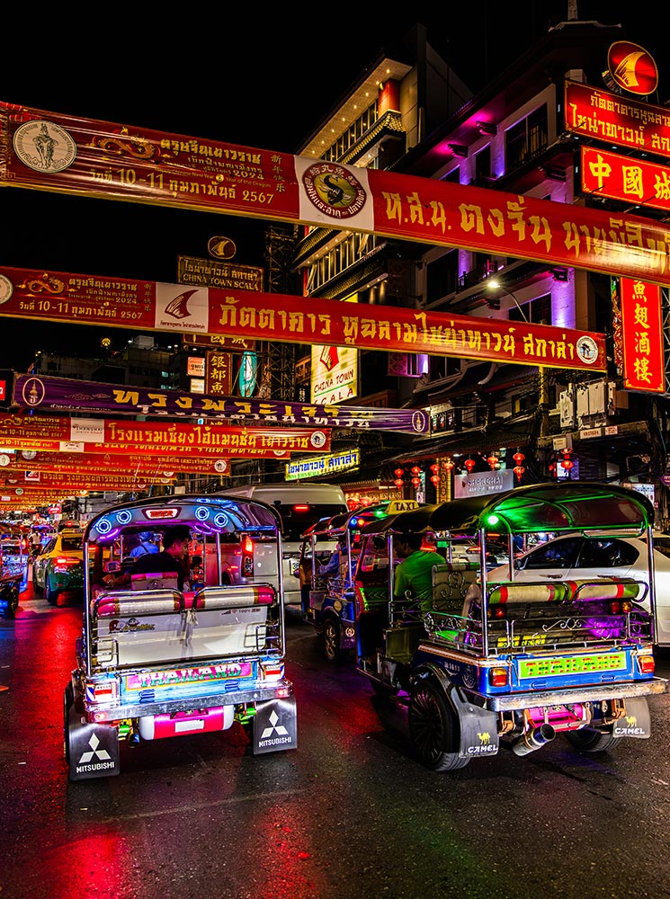 This image captures a vibrant nighttime scene in Bangkok's Chinatown, with colorful tuk-tuks lined up on a busy street illuminated by glowing neon lights and traditional red banners with Thai script. The bright reflections on the wet road add a dynamic feel to the bustling atmosphere, highlighting the energy and charm of Bangkok’s iconic tuk-tuk culture.