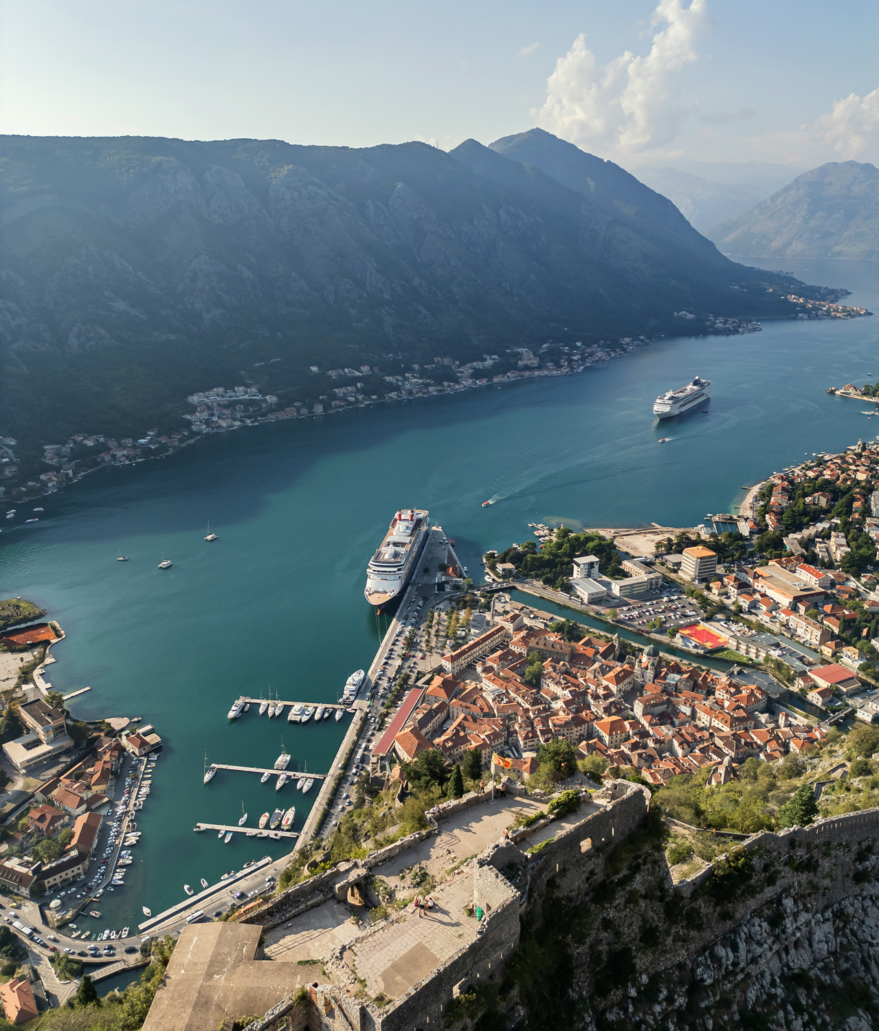 An aerial view of the Bay of Kotor showcasing the historic Old Town with its terracotta roofs, a cruise ship docked at the harbor, and dramatic mountains surrounding the serene blue water.