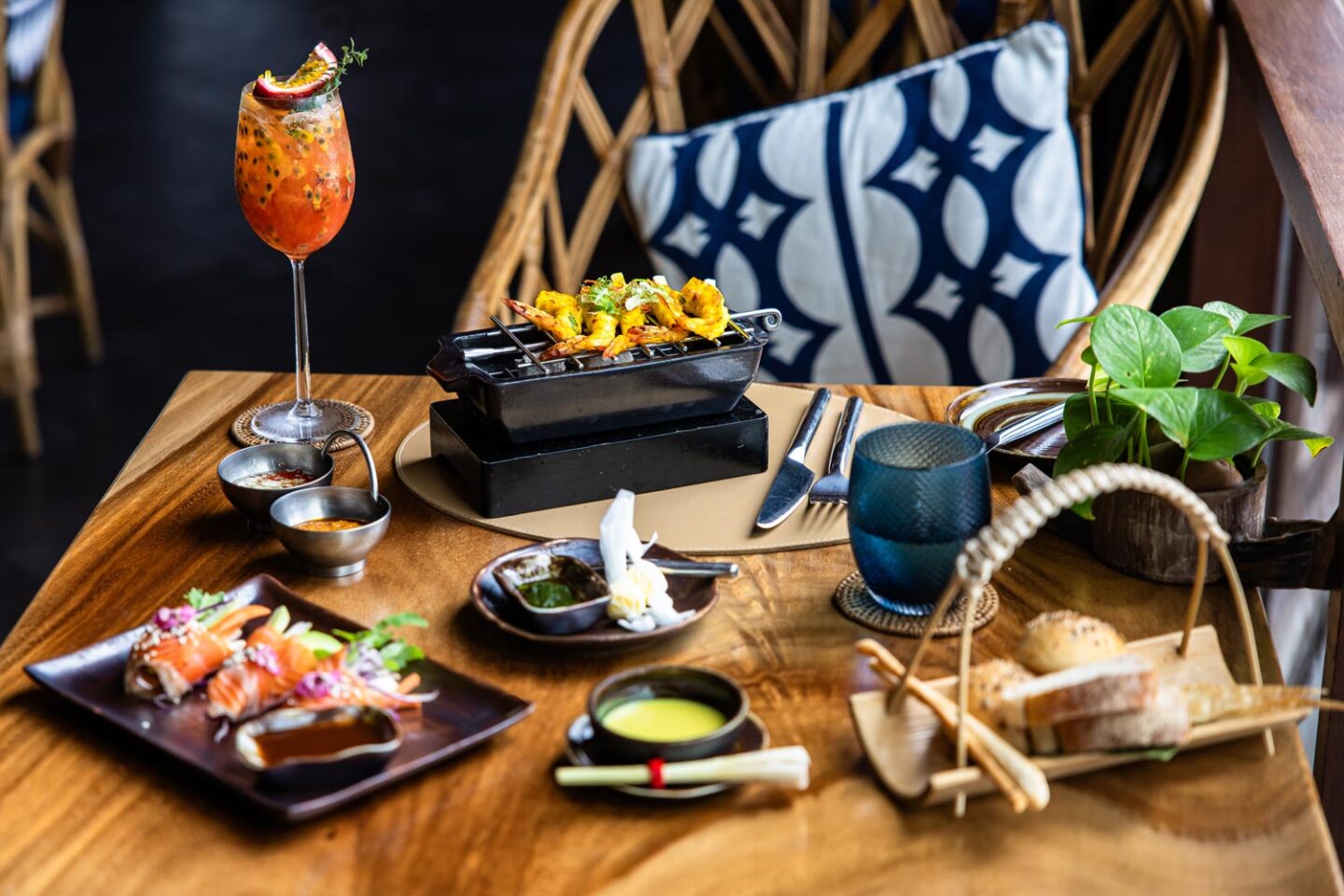 This image showcases an elegant dining setup featuring a variety of gourmet dishes and drinks on a polished wooden table. The spread includes grilled skewers served on a small tabletop grill, a plate of sushi-style appetizers, a basket of artisan bread, a bowl of soup, and a vibrant passion fruit cocktail. The background features cozy seating with blue and white patterned cushions, creating a refined yet inviting atmosphere. 