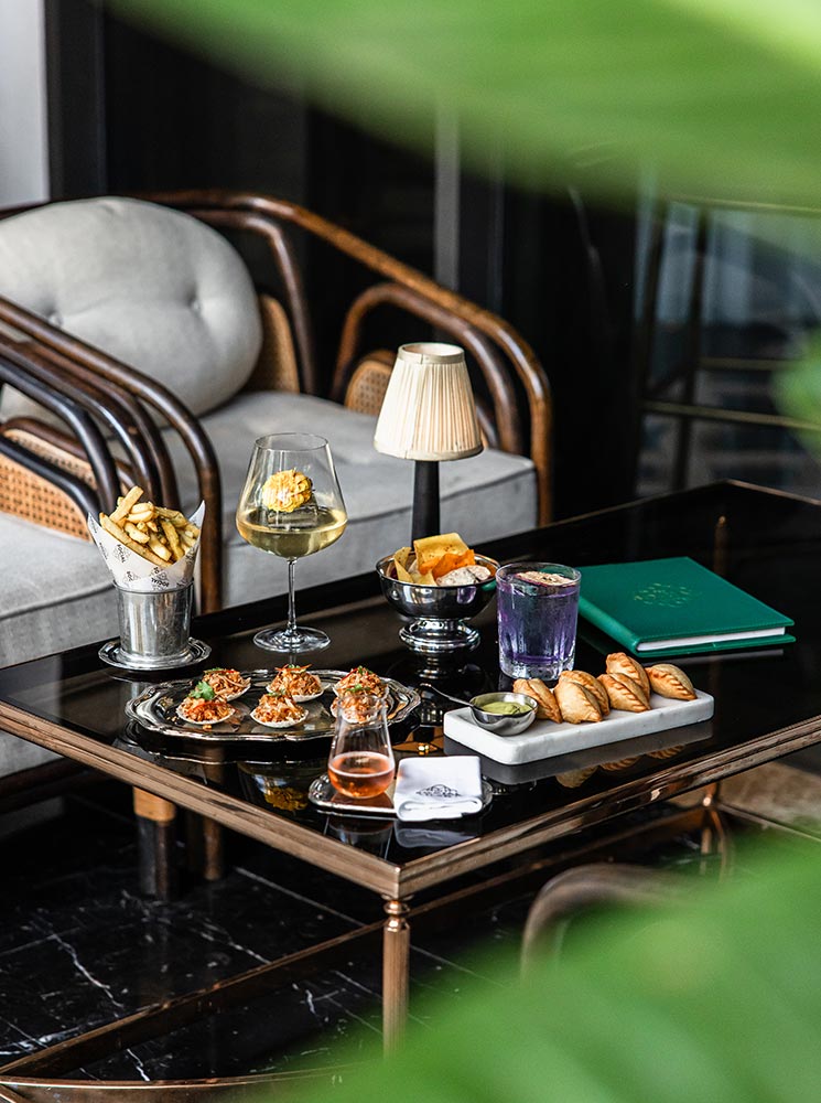 This image captures a chic and refined dining setup at the BKK Social Club in the Four Seasons Hotel Bangkok. The table features an array of elegant appetizers, including empanadas, fries, and bite-sized seafood dishes, paired with vibrant cocktails and wine. The sophisticated decor, complete with cozy seating and soft lighting, enhances the upscale ambiance.