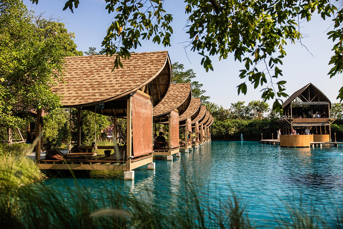 This image features a serene lakeside cafe with wooden pavilions built over turquoise water, offering a tranquil escape surrounded by greenery. The curved roofs of the structures and the lush foliage framing the scene create a harmonious and inviting atmosphere. A larger wooden building on stilts can be seen in the background, adding depth to this picturesque setting.