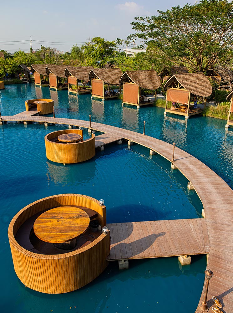 This image captures a serene dining area featuring wooden pavilions and circular seating platforms built over a vibrant blue pond. Connected by curved wooden walkways, the setup creates a tranquil and unique floating experience surrounded by lush greenery. The natural and modern elements blend seamlessly, offering an inviting and picturesque atmosphere.