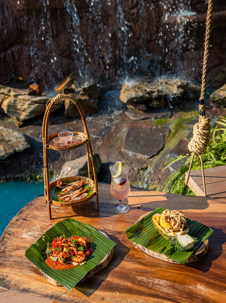 This image showcases a rustic dining setup featuring traditional Thai dishes presented on banana leaves, placed on a wooden table near a cascading waterfall. The dishes include spicy shrimp curry, pineapple fried rice, and grilled chicken, accompanied by a refreshing pink drink with a slice of lemon. The natural backdrop of rocks, water, and greenery creates a tranquil and inviting atmosphere.