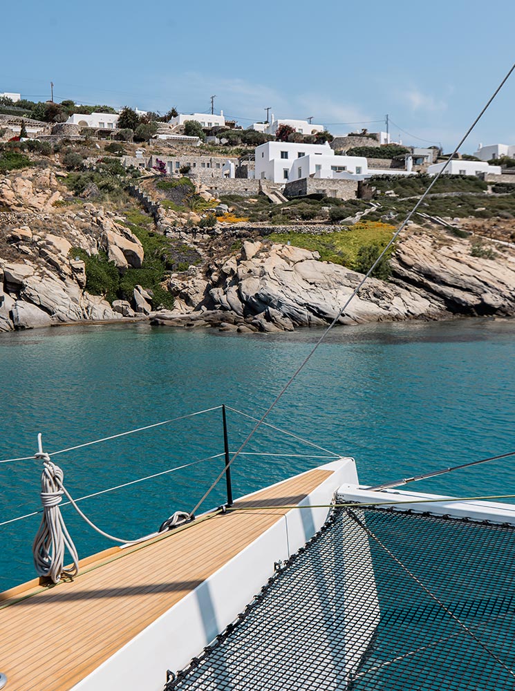 The deck of a catamaran overlooking turquoise waters near Mykonos, with whitewashed hillside homes and rocky terrain in the background under a clear blue sky.