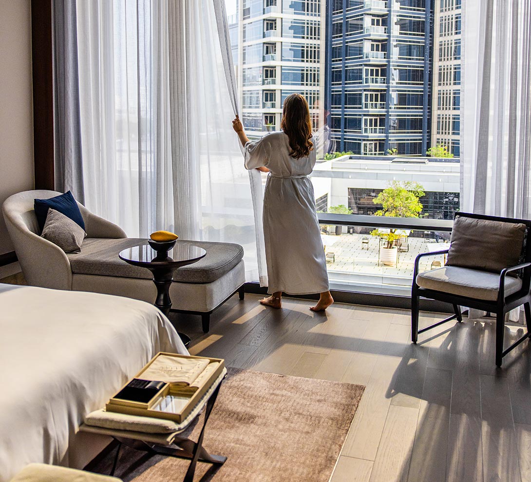 This image captures the interior of a luxurious deluxe room at the Four Seasons Hotel Bangkok, featuring elegant furnishings and floor-to-ceiling windows. A woman in a white robe gazes out at the cityscape and modern buildings, creating a serene and sophisticated atmosphere. The room includes a plush bed, a chaise lounge, and a stylish table with decor, emphasizing comfort and refinement.