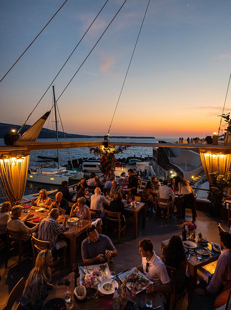 An evening dining scene at Ammoudi Fish Tavern in Santorini, with guests enjoying meals under warm lighting, overlooking the sea and a vibrant sunset sky, surrounded by sailboats and waterfront views.