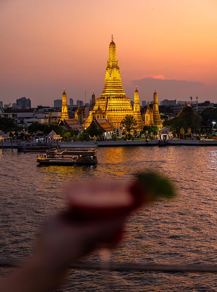 This image features a breathtaking view of Wat Arun, the Temple of Dawn, glowing warmly at sunset on the banks of the Chao Phraya River. In the foreground, a blurred hand holds a red cocktail garnished with lime, adding a modern rooftop bar vibe to the serene and iconic scene. The soft orange and pink hues of the sky enhance the tranquil atmosphere.