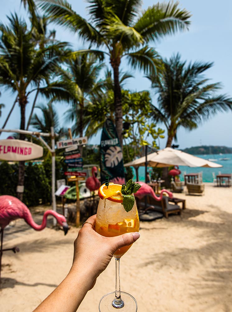 A hand holding a tropical cocktail garnished with orange slices and mint, set against a beach scene with pink flamingo decorations, palm trees, and seating overlooking the turquoise ocean.