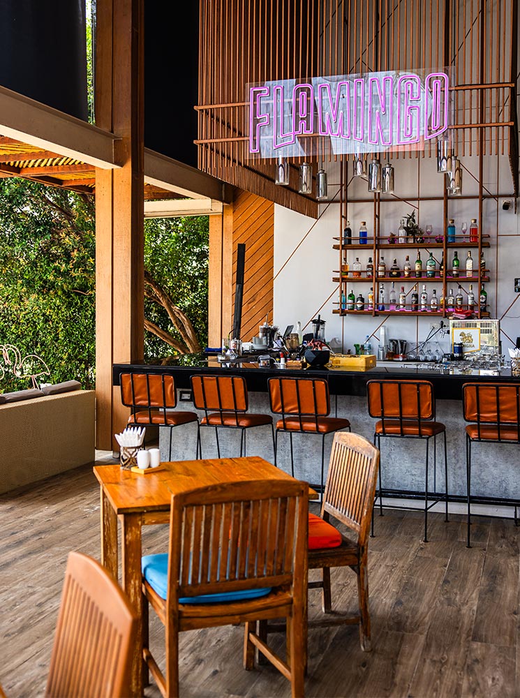 Interior of a beach bar with wooden tables and chairs, a bar counter lined with leather stools, and a neon pink "Flamingo" sign above shelves stocked with bottles, creating a tropical and stylish ambiance.