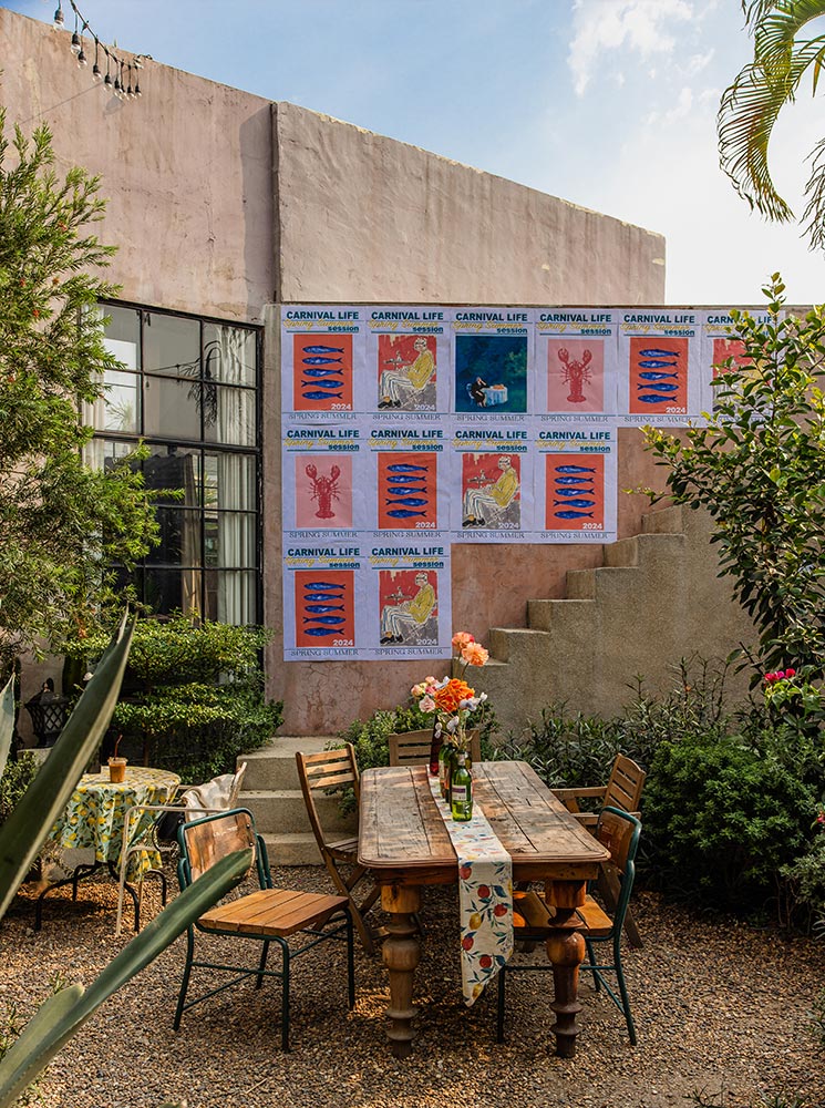 This image shows a cozy outdoor cafe setting with a rustic wooden table and chairs arranged on a gravel courtyard. The table is decorated with a floral table runner and fresh flower vases, while colorful posters titled "Carnival Life" adorn the stucco wall in the background. The lush greenery and pastel-toned architecture create a relaxed and inviting atmosphere.