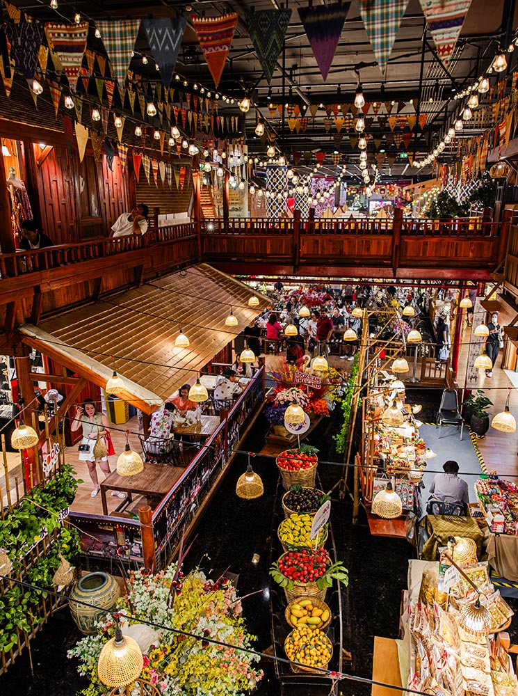 This image captures a lively floating market scene, with rows of vibrant fruit and flower displays in woven baskets arranged on wooden boats. The market is surrounded by a traditional wooden structure, adorned with colorful pennant flags and hanging lights, adding to the festive atmosphere. Visitors can be seen exploring the bustling space, reminiscent of Thailand’s cultural and culinary heritage.