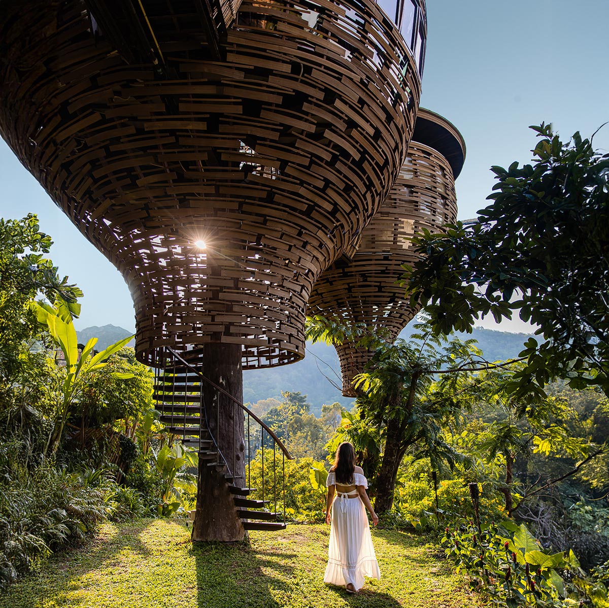 This image features a stunning view of the Keemala Resort in Phuket, showcasing its iconic woven pod-like treehouses elevated above lush greenery. A woman in a flowing white dress walks toward a spiral staircase leading up to one of the structures, with sunlight streaming through the intricate wooden design. The scene evokes a sense of tranquility, luxury, and harmony with nature. 







