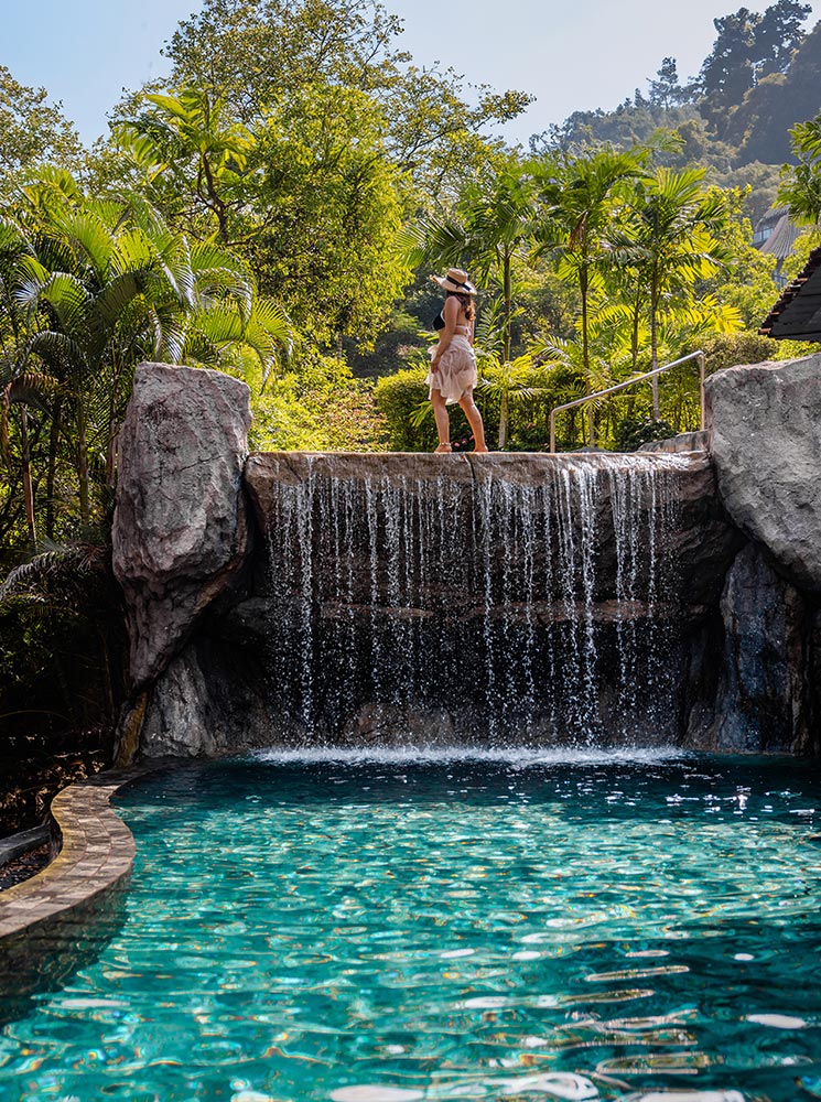 This image showcases a serene pool at the Keemala Resort in Phuket, with a cascading waterfall flowing from a rocky ledge into crystal-clear turquoise water. A woman dressed in a light outfit and sunhat stands atop the waterfall, surrounded by lush tropical greenery under a bright sky. The scene exudes relaxation and harmony with nature, highlighting a luxurious and tranquil escape.