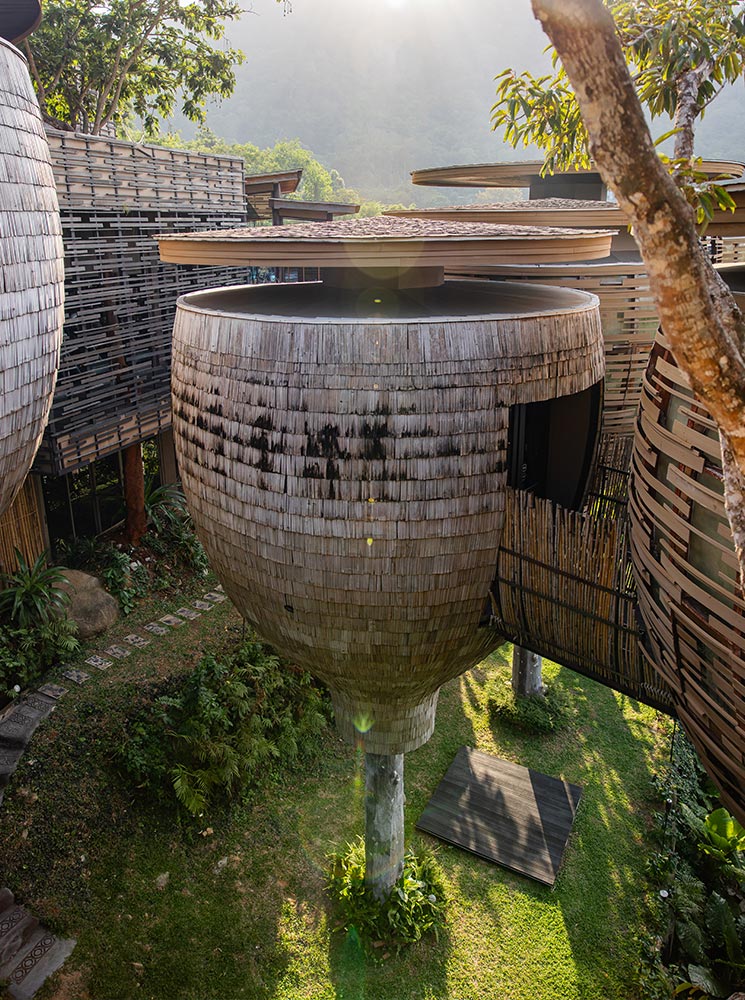 This image showcases one of the elevated pod-like villas at Keemala Resort in Phuket, designed with a unique woven exterior that blends seamlessly into the lush natural surroundings. The villa is supported by a single column, rising above a landscaped garden with soft sunlight filtering through the trees. The architectural design emphasizes harmony with nature and an eco-luxury aesthetic.