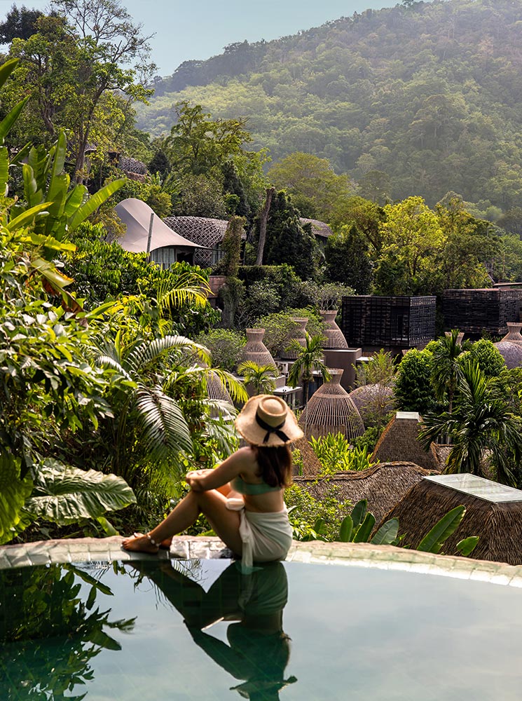 This image features a serene view of a woman sitting on the edge of an infinity pool overlooking the lush greenery and unique pod-like villas of Keemala Resort in Phuket. The surrounding landscape is rich with tropical plants and rolling hills, creating a tranquil and luxurious atmosphere. The architectural harmony with nature enhances the sense of escape and relaxation.