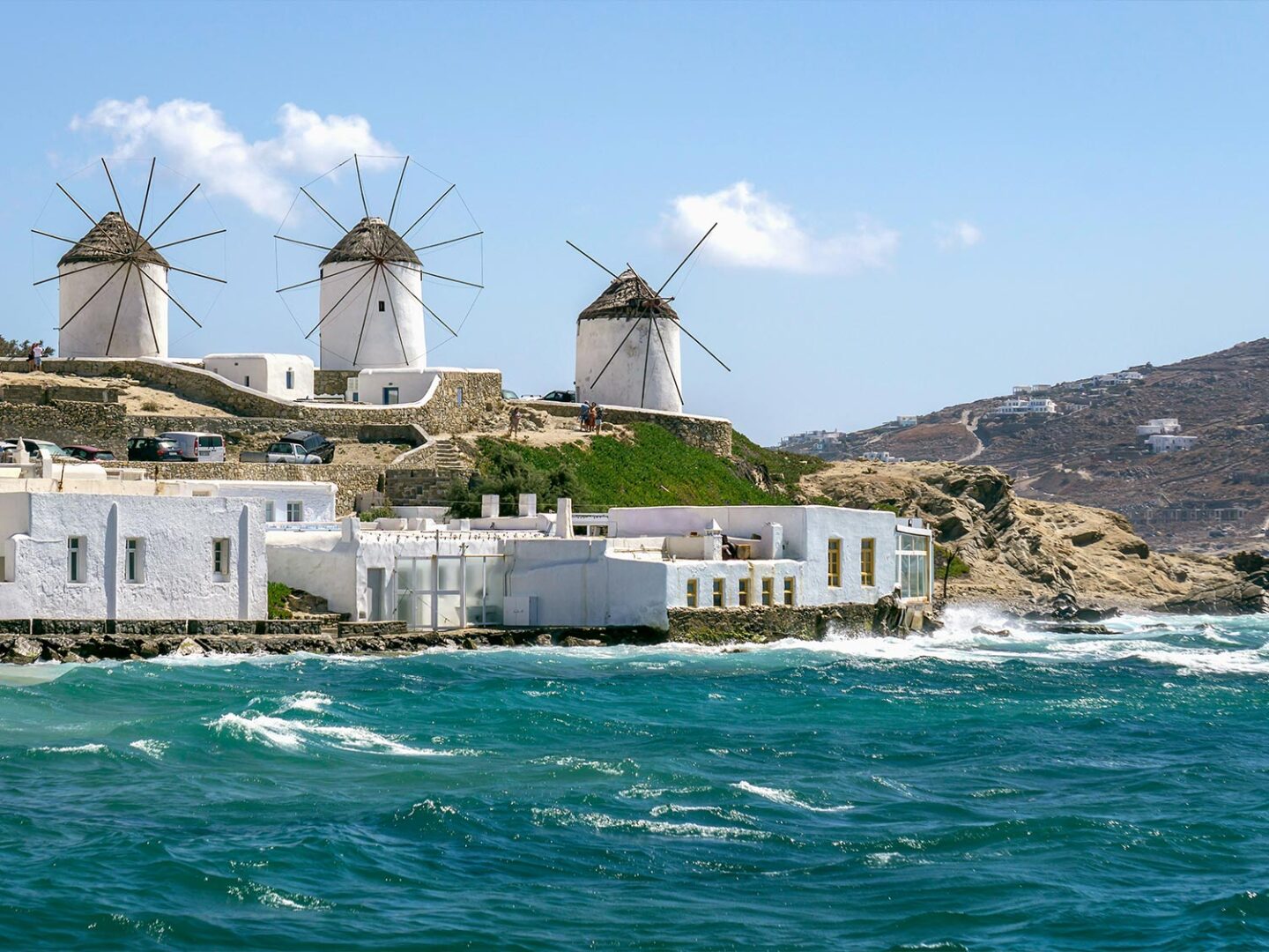 The iconic Kato Mili windmills of Mykonos, Greece, perched on a hill overlooking the turquoise Aegean Sea. Traditional whitewashed buildings and rocky coastline complete the scenic view under a clear blue sky.