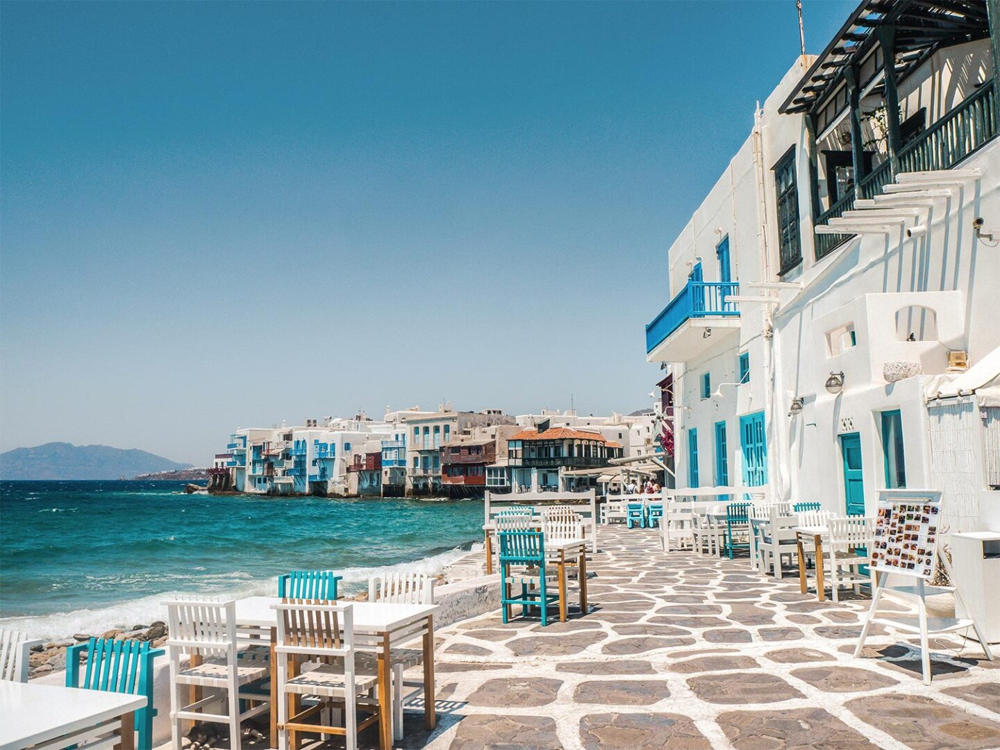 A picturesque seaside promenade in Mykonos, Greece, featuring whitewashed buildings with blue accents overlooking the Aegean Sea. Outdoor seating with white and turquoise chairs lines the cobblestone pathway, with waves gently lapping the shoreline.