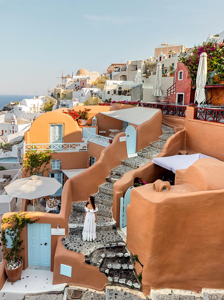 A charming view of terracotta-colored houses in Oia, Santorini, with curved stone steps, vibrant flowers, and whitewashed buildings in the background, featuring a woman in a white dress admiring the scenery.