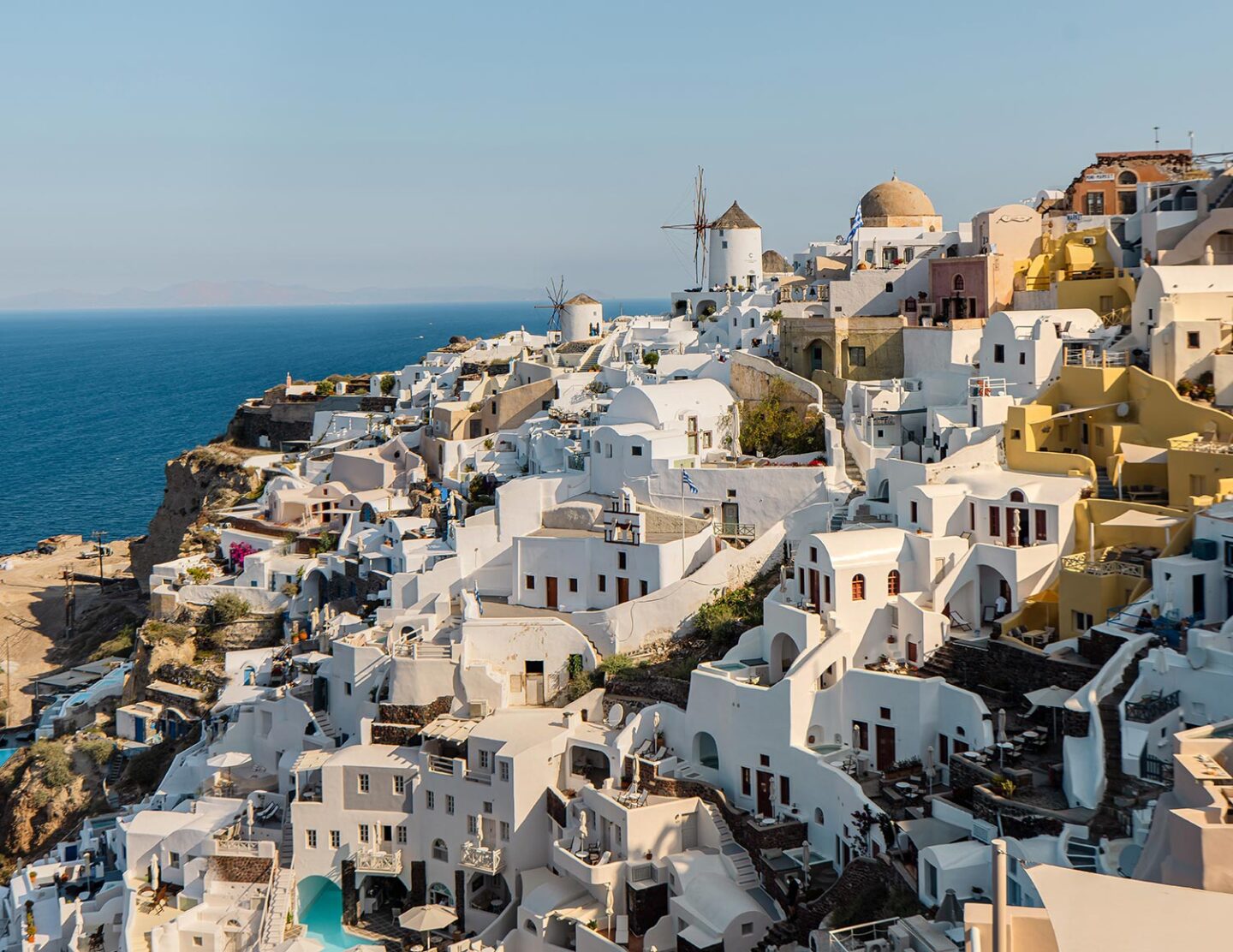 A stunning view of Oia, Santorini, featuring whitewashed buildings, domed roofs, and a traditional windmill perched on a cliff, overlooking the deep blue Aegean Sea under a clear sky.