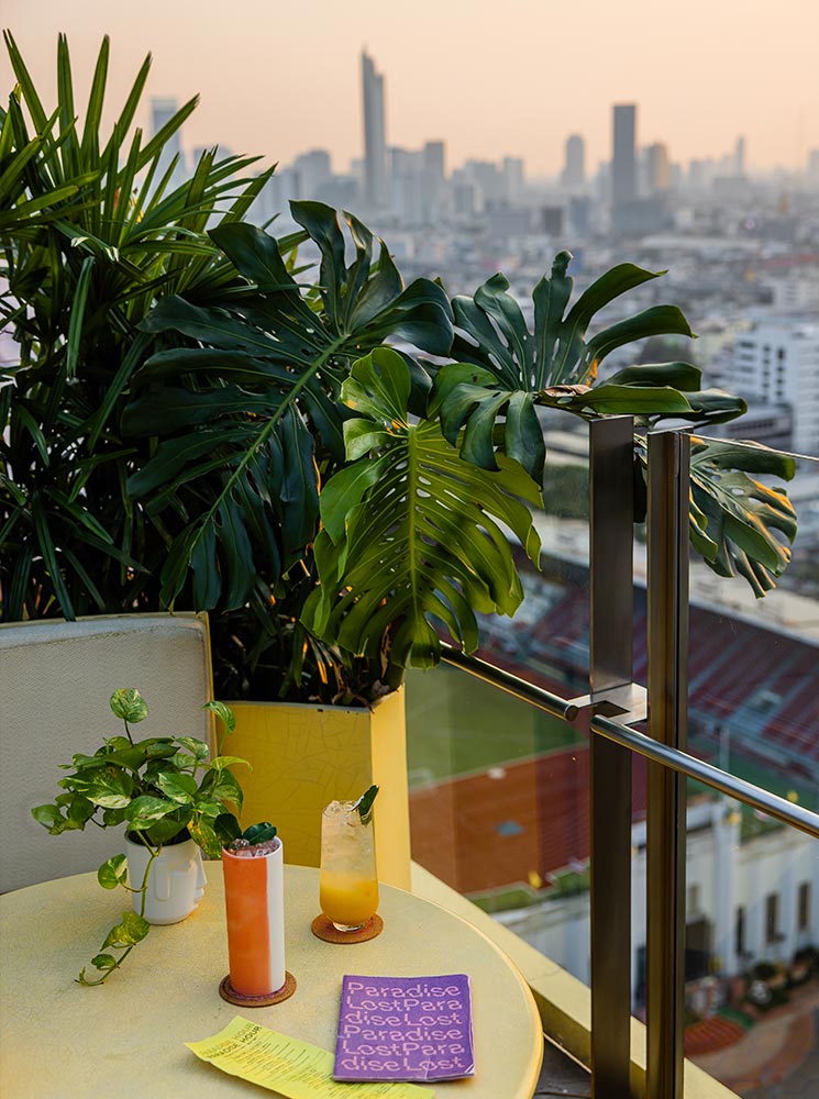 This image showcases a rooftop bar setting with a small round table featuring two colorful cocktails, potted plants, and a purple menu titled "Paradise Lost." The backdrop reveals a hazy Bangkok skyline during sunset, with tall buildings and a stadium visible in the distance. The lush green foliage framing the scene adds a tropical touch to the urban ambiance.