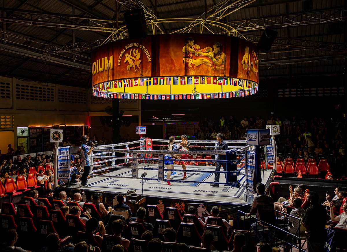 Muay Thai match in progress at Patong Boxing Stadium in Phuket, Thailand, with fighters in the ring, spectators in red stadium seats, and a brightly lit circular banner displaying images of fighters and international flags above the ring.