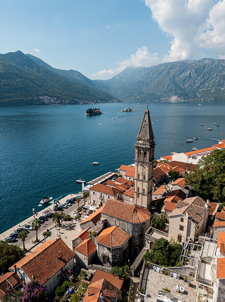 A scenic view of Perast in the Bay of Kotor, Montenegro, featuring terracotta-roofed historic buildings, a tall church bell tower, and calm blue waters with small islands and surrounding mountains in the background.