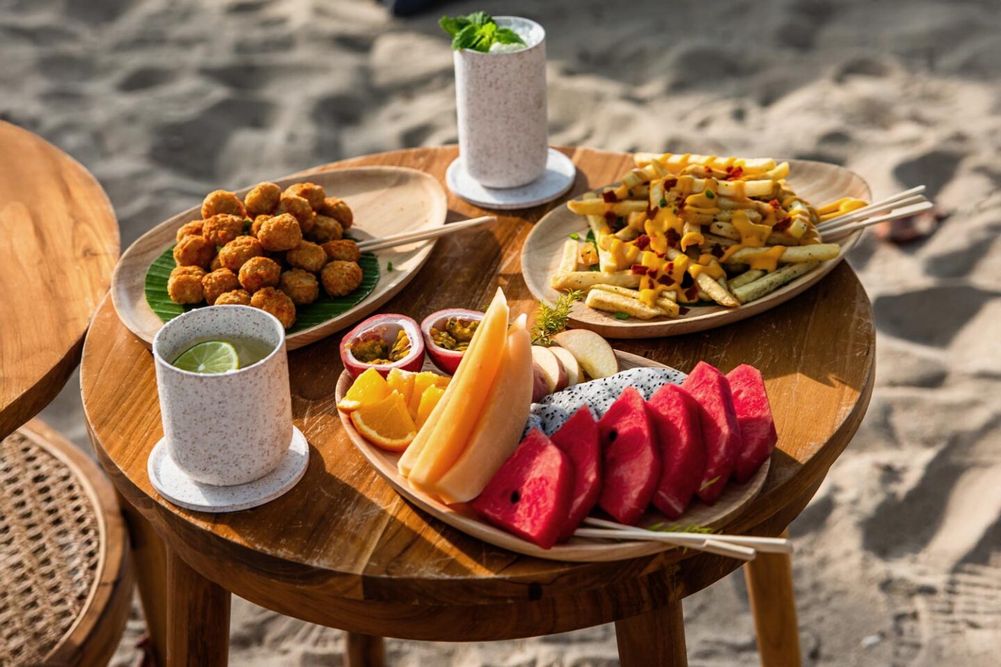 This image features a casual beachside dining setup with wooden tables displaying a variety of snacks and drinks. The spread includes fresh tropical fruits like watermelon, dragon fruit, orange slices, and passion fruit, along with crispy fried bites and fries topped with cheese and seasonings. Two refreshing drinks, garnished with lime and mint, complete the inviting and relaxed atmosphere, with sandy beach surroundings in the background.