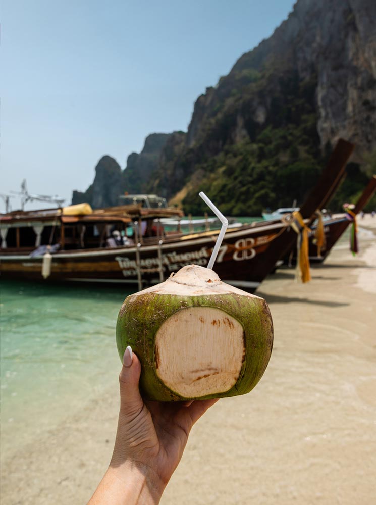 This image features a hand holding a freshly cut coconut with a straw, set against the backdrop of a sandy beach and clear turquoise water. Traditional Thai long-tail boats are docked on the shore, with towering limestone cliffs visible in the distance. The composition captures a quintessential tropical vibe, emphasizing relaxation and natural beauty.