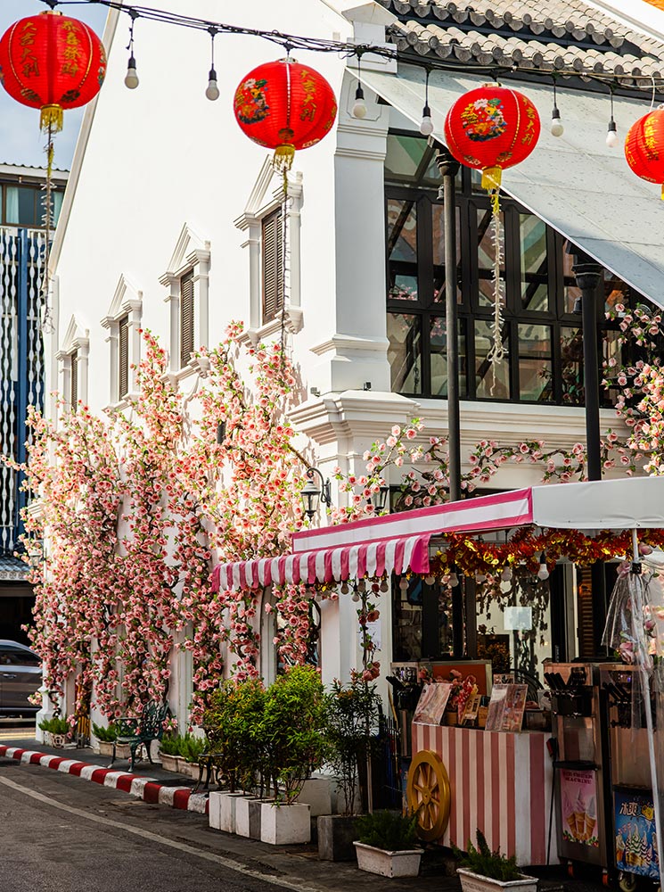 This image showcases a picturesque street scene in Phuket Old Town, featuring a white colonial-style building adorned with an abundance of pink and white floral decorations. Red lanterns hang across the street, adding a festive ambiance, while a striped pink-and-white awning shelters a charming storefront offering drinks or snacks. The setting exudes a blend of traditional and romantic charm, perfect for a leisurely stroll or photo opportunity.