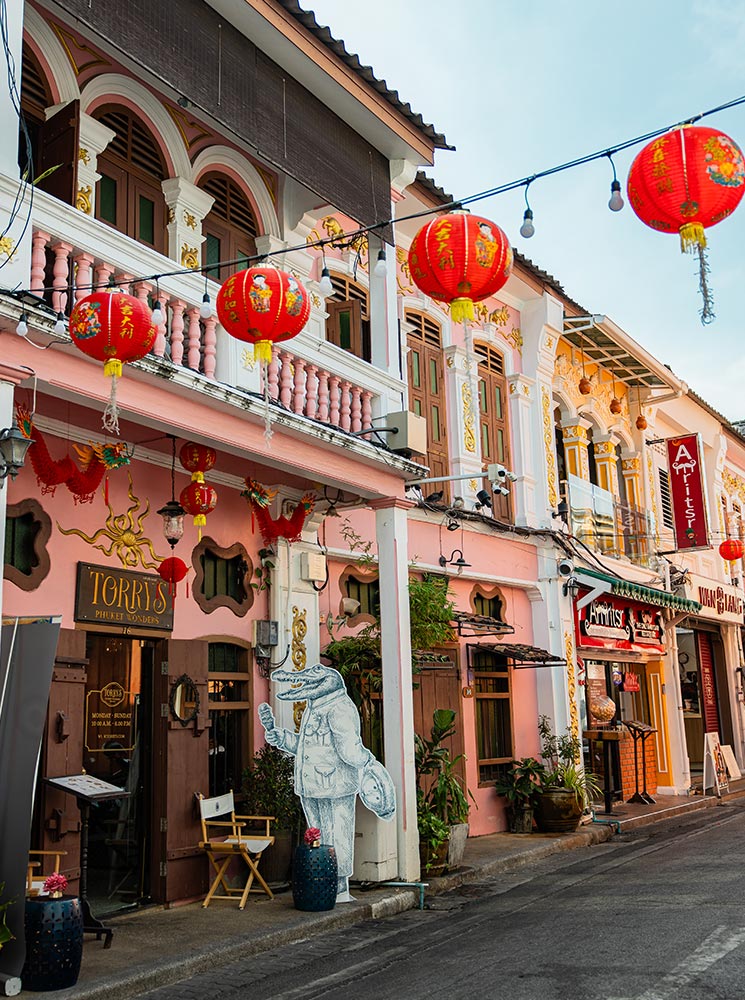 This image captures a lively street scene in Phuket Old Town, featuring pastel-colored Sino-Portuguese buildings adorned with intricate details. Red lanterns hang overhead, adding a festive and traditional touch, while the storefronts, including one with a crocodile figure art display, reflect a mix of heritage and contemporary charm. The vibrant atmosphere highlights the area's cultural and architectural significance.