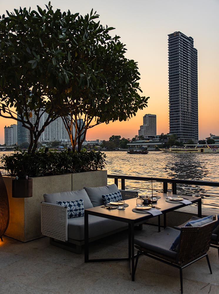 This image showcases an elegant outdoor dining area overlooking the Chao Phraya River during sunset at the Four Seasons Hotel Bangkok. The table is set with minimalist tableware, surrounded by comfortable seating with decorative cushions, and framed by lush greenery. The calm river and towering cityscape in the background add to the sophisticated and tranquil atmosphere.