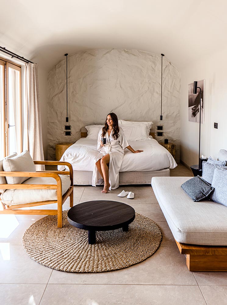 A stylish hotel room with a cozy bed framed by textured white walls, a wooden armchair, a woven circular rug, and modern minimalist decor, featuring a woman in a white robe sitting on the bed holding a cup.