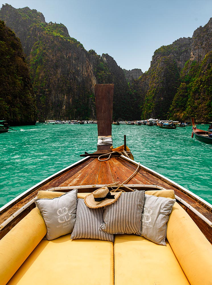 This image depicts the view from a traditional long-tail boat as it glides through turquoise waters, surrounded by towering limestone cliffs at a popular destination like the Phi Phi Islands in Thailand. The boat features cozy yellow seating adorned with striped and branded cushions, along with a straw hat, emphasizing a relaxed and scenic adventure. The stunning backdrop of clear waters and dramatic cliffs enhances the tropical paradise vibe.