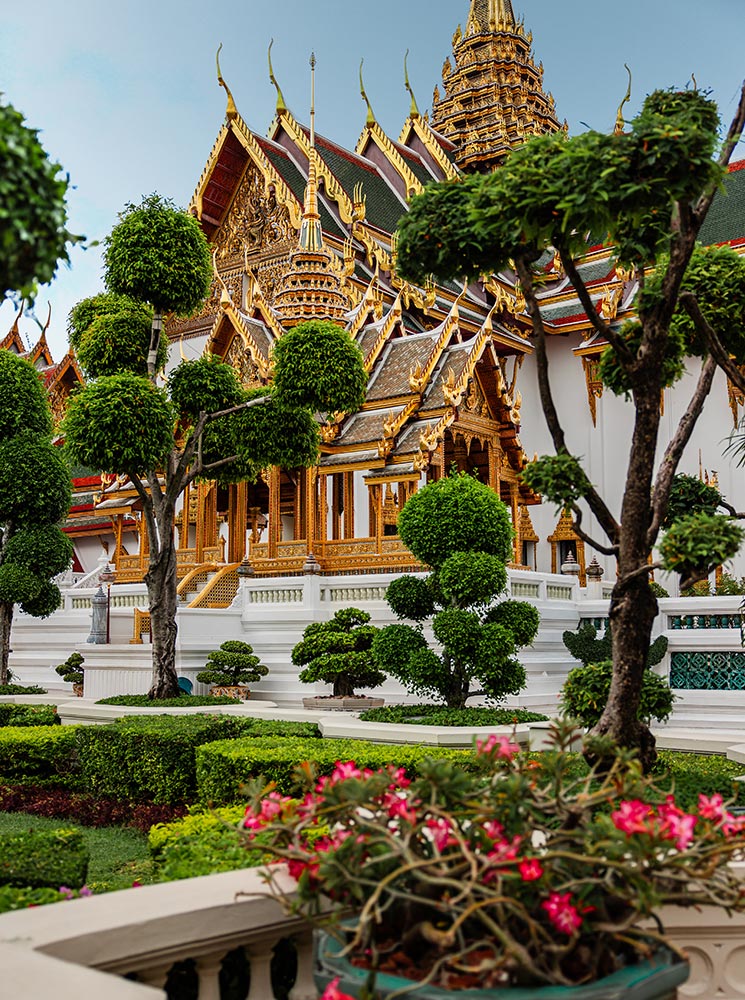 This image showcases the ornate architecture of the Grand Palace in Bangkok, Thailand, with its golden spires, intricate carvings, and traditional Thai roof design. The foreground features manicured gardens with neatly trimmed trees and vibrant pink flowers, enhancing the elegance of the palace’s grand design.