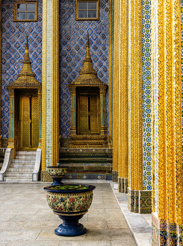 This image highlights the intricate design of the Grand Palace in Bangkok, with golden columns adorned with ornate mosaic patterns set against a rich blue tiled wall. The scene features a decorative ceramic planter in the foreground, adding depth and contrast to the grandeur of the palace's detailed craftsmanship.